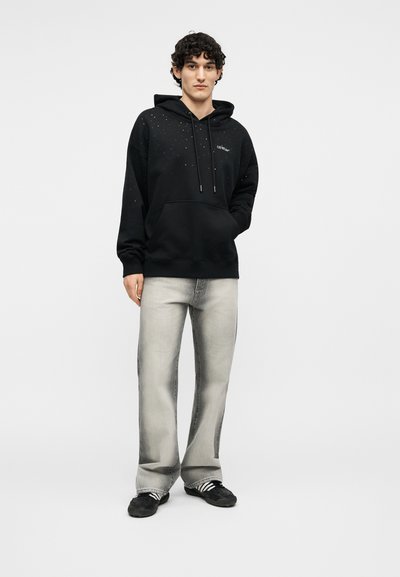 Jauns vīrietis ar cirtainiem matiem, tērpies melnā kapučjakā ar plankumiem, gaiši pelēnās džinsa biksēs un melnās sporta kurpēs ar baltām svītrām, stāv pie vienkārša fona.