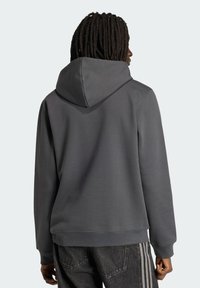 Άτομο με σκούρες κοτσίδες, που φοράει απλό γκρι σκούρο φούτερ με κουκούλα και μαύρα τζιν με ανοιχτές ρίγες, γυρισμένο προς τα πίσω, μπροστά σε φωτεινό φόντο.