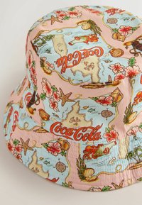Chapeau bob avec carte vintage colorée, créatures marines, fleurs et motifs du logo "Coca-Cola" sur fond rose clair et bleu.