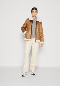 Giubbotto in shearling sintetico marrone con colletto e polsini di pelliccia bianca, abbinato a pantaloni crema chiari e sneakers bianche sportive.
