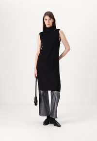 Μαύρο αμάνικο φόρεμα με ψηλή λαιμόκοψη, συνδυασμένο με μεταλλικά παντελόνια σε φαρδιά γραμμή και μαύρα παπούτσια, κρατώντας μια μικρή τσάντα.