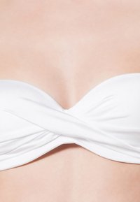 Bijeli bikini top bandeau s dizajnom uvrnutog prednjeg dijela, izrađen od glatkog materijala, s bešavnom teksturom i minimalističkim stilom.