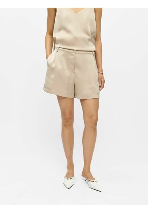 Frau trägt beige ärmelloses Oberteil und passende hochtaillierte Shorts, steht mit Händen teilweise in den Taschen der Shorts, weiße spitze Schuhe.