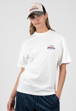 Kvinde iført hvid T-shirt med teksten "Prohibited Sporting Goods Inc." og matchende beige og sort kasket, kombineret med marineblå stribede bukser, står afslappet.