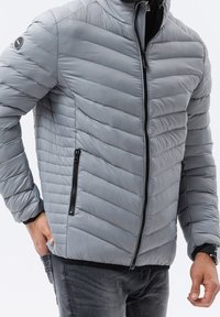 Szara kurtka puchowa z wzorem w jodełkę, wysokim kołnierzem i zamkiem błyskawicznym z przodu. Posiada czarne kieszenie na zamek i miękką, gładką teksturę.