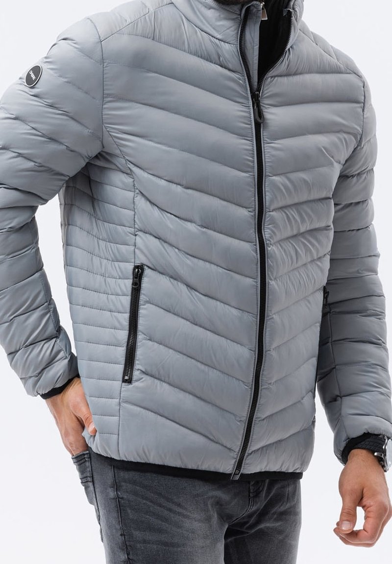 Szara kurtka puchowa z wzorem w jodełkę, wysokim kołnierzem i zamkiem błyskawicznym z przodu. Posiada czarne kieszenie na zamek i miękką, gładką teksturę.