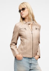 Γυναίκα με μπεζ δερμάτινο μπουφάν με φερμουάρ σε τσέπες και ανοιχτό μπλε τζιν, με τα χέρια στις τσέπες, κοιτάζοντας πλάγια, φορώντας γυαλιά ηλίου.