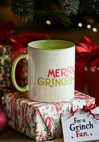 Tazza in ceramica con interno e manico verdi, esterno bianco con la scritta "MERRY GRINCH" in rosso e giallo. Circondata da regali e decorazioni natalizie.