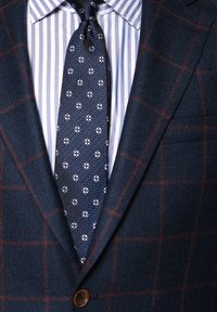 Granatowa marynarka w kratę z brązowymi akcentami noszona na jasnoniebieskiej koszuli w białe paski, uzupełniona ciemnym krawatem w kwiatowy wzór.