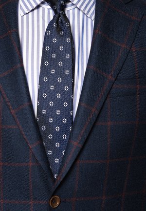 Granatowa marynarka w kratę z brązowymi akcentami noszona na jasnoniebieskiej koszuli w białe paski, uzupełniona ciemnym krawatem w kwiatowy wzór.