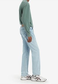 Svetle modré džíny s rovnými nohavicemi kombinované s dlouhým zeleným tričkem, doplněné bílými teniskami s černými a červenými akcenty.