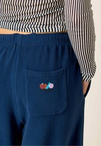 Pantalon bleu marine avec une texture douce, comportant une poche arrière ornée d'un design de broderie de fruits colorés. Chemise rayée visible au-dessus.