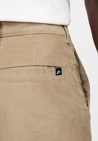 Pantalones de pana beige con un corte regular, superficie texturizada y una pequeña etiqueta negra del logo de Nike en el bolsillo trasero.