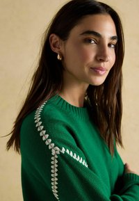 Grüner gestrickter Pullover mit geripptem Kragen und einzigartiger weißer Naht entlang der Schulternähte. Weicher Stoff mit lässiger Passform.