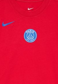 T-shirt en coton rouge avec un logo Nike bleu en haut à gauche et un emblème circulaire bleu du Paris Saint-Germain affiché au centre.