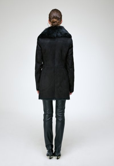 Femme avec les cheveux en chignon portant un manteau noir à col en fourrure, un pantalon en cuir noir et des talons noirs, debout de dos sur un fond uni.