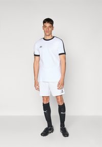 Balts sporta krekls ar melniem akcentiem, kopā ar baltām šortiem. Modelis valkā melnas futbola zeķes un sporta apavus, uzsverot sportisko dizainu.