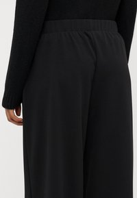 Pantalon large noir avec une ceinture élastique lisse et un tissu doux et texturé. Le design présente une coupe ample et droite.