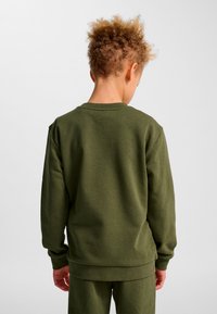 Barn med lockigt hår som bär en olivgrön sweatshirt och matchande byxor, stående med ryggen mot en enkel bakgrund.