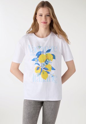 Giovane donna con t-shirt bianca decorata con limoni gialli e foglie azzurre, abbinata a jeans grigi, in piedi davanti a uno sfondo bianco.