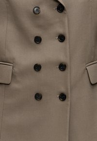 Gros plan d'un manteau beige croisé avec six boutons foncés et deux poches à rabat de chaque côté.