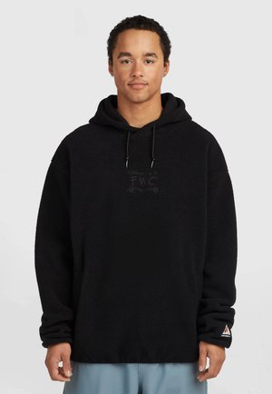 Jonge man die een zwarte fleecehoodie draagt met een subtiel logo op de borst en linker mouw, staand tegen een effen lichte achtergrond.