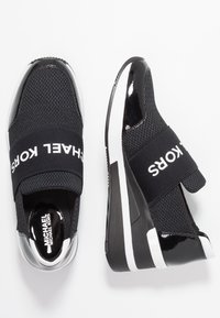 Schwarze Sneakers aus Mesh mit einem doppelten Riemen und weißem Schriftzug. Glänzende schwarze Akzente und eine strukturierte Sohle prägen das Design.