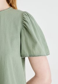 Svetlozelena srajca z kratkimi nabranimi rokavi in valovitimi robovi. Mehka tekstura in sproščen kroj.