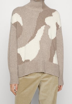 Pull tricoté beige et crème avec un motif abstrait, col montant et poignets côtelés, porté avec un pantalon beige en velours côtelé.