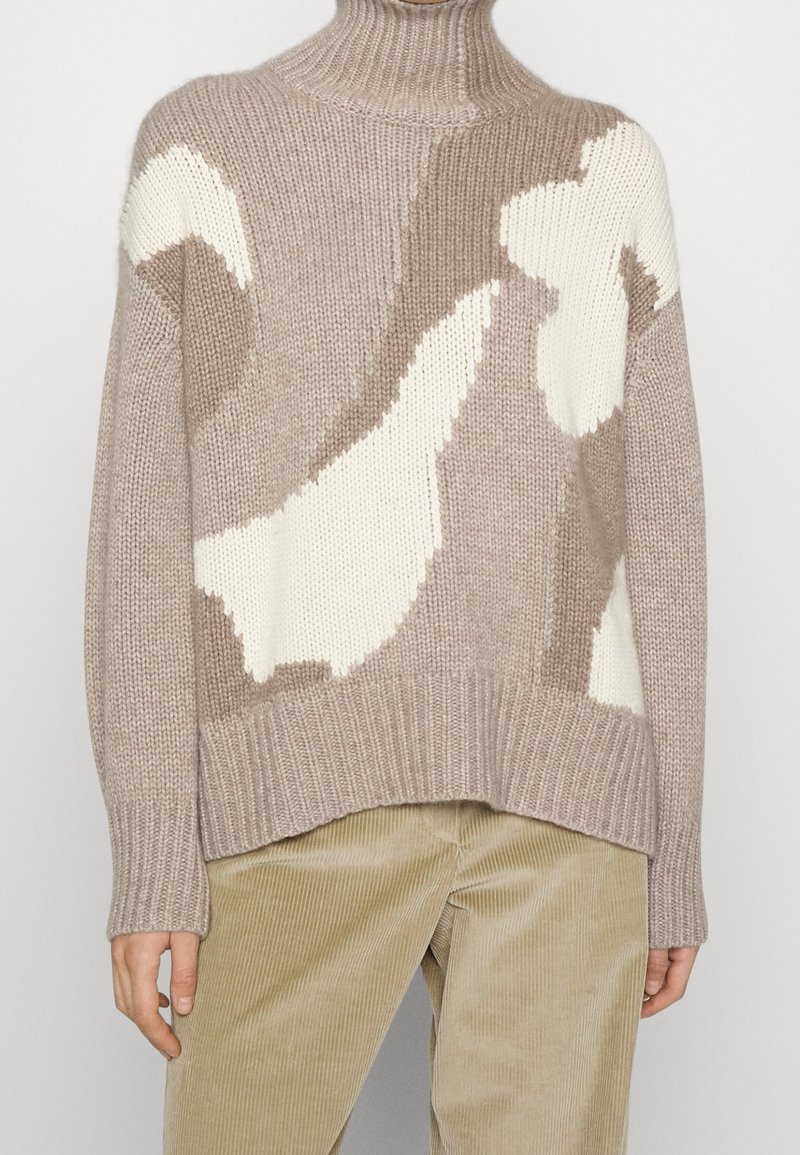 Pull tricoté beige et crème avec un motif abstrait, col montant et poignets côtelés, porté avec un pantalon beige en velours côtelé.