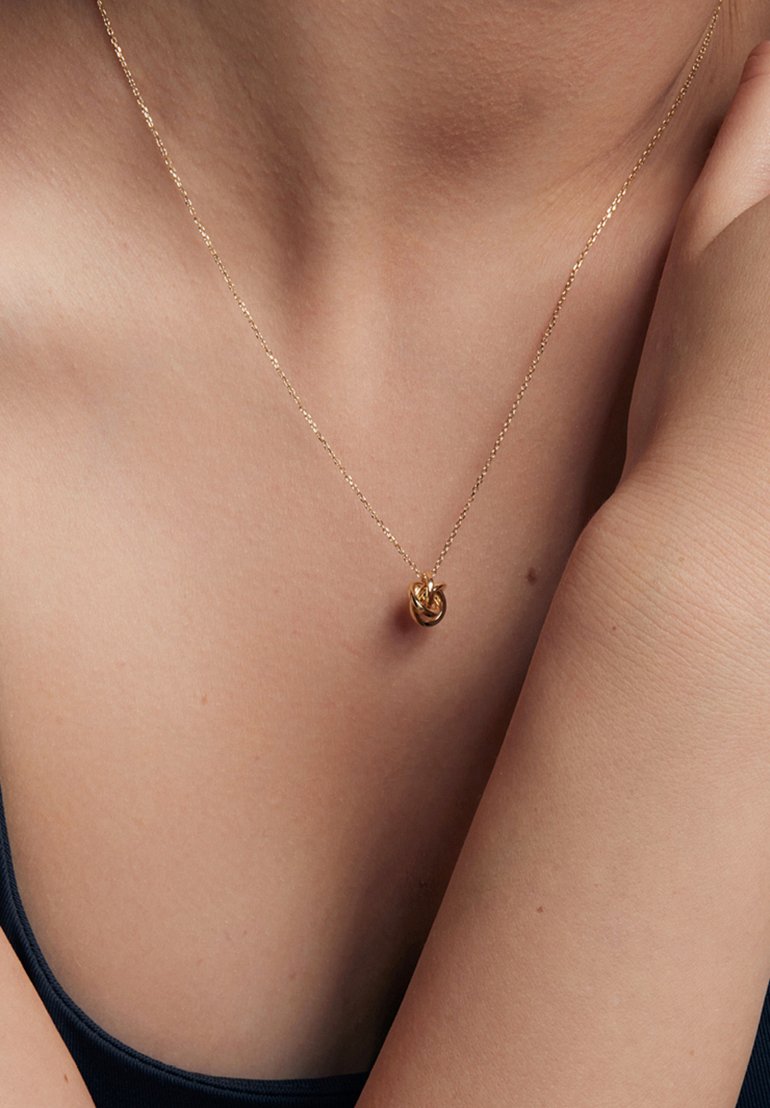 Złoty naszyjnik z skręconym wisiorkiem, delikatnie wplecionym łańcuchem i gładką fakturą, noszony na nagiej skórze. Subtelny blask z minimalnymi detalami projektu.