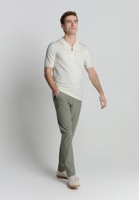 Crème polo met korte mouwen en ritssluiting, gecombineerd met lichtgroene op maat gemaakte broek en beige sneakers, lopend tegen een neutrale achtergrond.