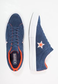 Zapatillas de ante azul marino con suelas de goma blanca. Presentan acentos rojos, cinco ojales y un logo en forma de estrella blanca en el lateral.
