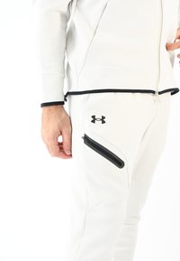 Chándal gris claro con un bolsillo con cremallera. Presenta una cremallera negra y detalles en negro, un diseño elegante y una tela texturizada para mayor comodidad.
