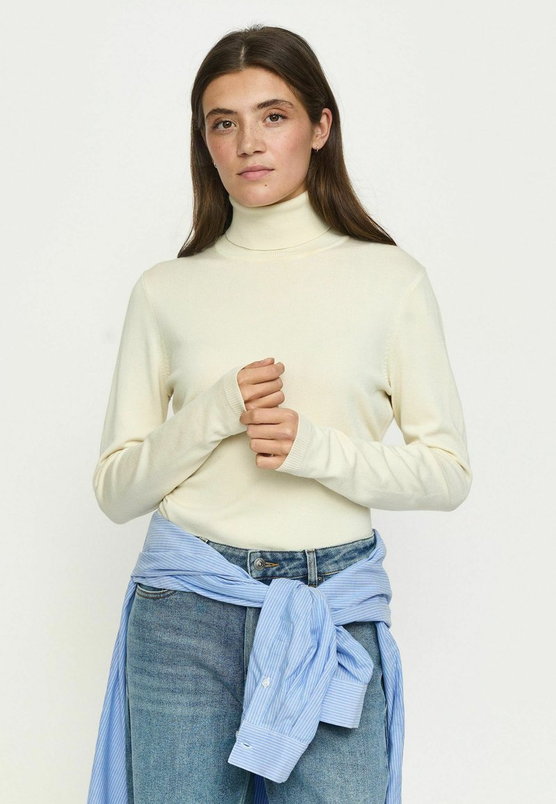 Jeune femme aux longs cheveux bruns portant un col roulé crème et un jean bleu avec une chemise bleue nouée autour de la taille.