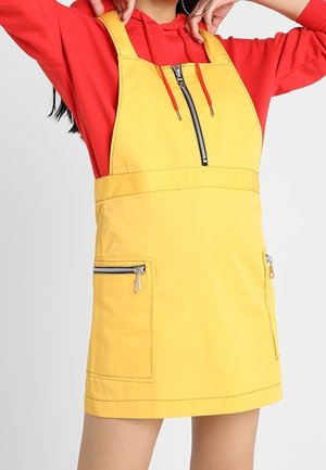Robe salopette jaune sans manches avec fermeture éclair à l'avant et poches zippées portée sur un sweat à capuche rouge.