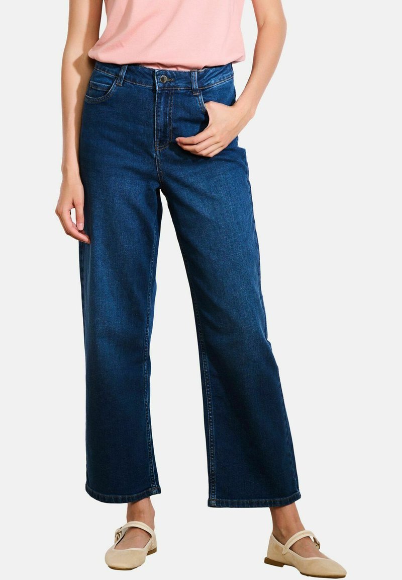 Jeans a gamba larga di colore blu scuro, a vita alta con tasche, realizzati in tessuto denim. Indossati con una camicia rosa chiaro e scarpe beige.