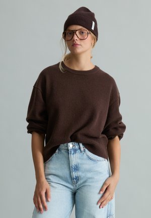 Vrouw die een bruine muts, bril en losse bruine trui draagt, gestopt in lichtblauwe high-waisted jeans tegen een effen grijze achtergrond.