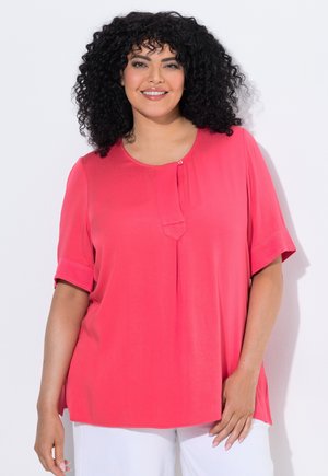 Lachende vrouw met krullend haar, gekleed in een felroze blouse met korte mouwen en witte broek, staand tegen een witte achtergrond.