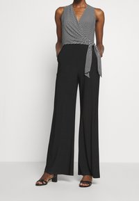 Svart vida byxdress med v-ringning, mönstrad halterneck-top och sidoknytning. Slätt tyg med fickor för extra funktionalitet.