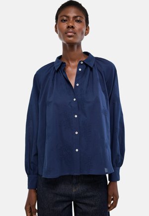 Femme portant un chemisier ample bleu marine à boutons avec des motifs floraux texturés subtils et des manches longues, associé à un jean foncé.