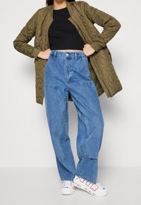 Persoon in een olijfgroene gewatteerde jas, zwarte cropped top, hoge, wijde blauwe spijkerbroek en witte sneakers met rode en blauwe details.