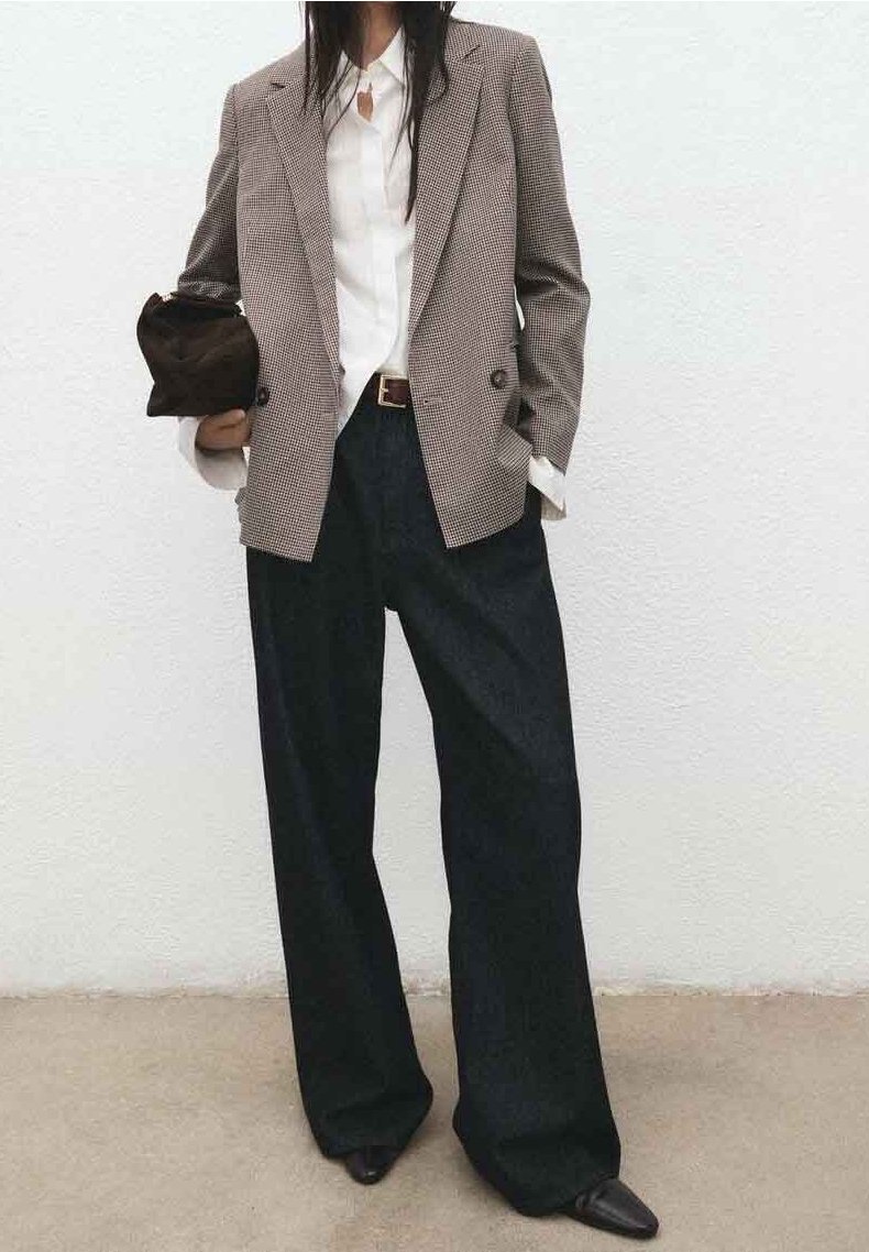 Femme portant un blazer marron à carreaux, une chemise blanche, un pantalon noir à jambes larges, des chaussures noires à bouts pointus, tenant une petite pochette noire.