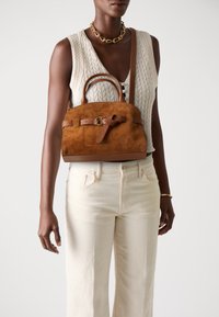 Brown suede handbag with leather accents, round handles, and a tied detail. Model wears a knit top and beige wide-leg pants.