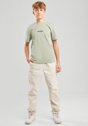 Light green cotton t-shirt with "INTO THE WILD" print, paired with cream-colored pants and white sneakers. Simple design, relaxed fit.