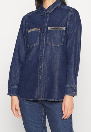 Vrouw draagt een donkerblauw denim overhemd met knopen en twee borstzakken, gecombineerd met een bijpassende blauwe spijkerbroek, staand tegen een witte achtergrond.