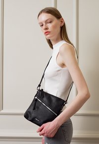 Černá kožená crossbody taška s texturovaným povrchem, vybavená přední kapsou na zip a nastavitelným popruhem. Jednoduchý, obdélníkový design.