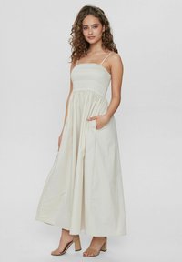 Maxikleid in hellem Creme mit gerafftem Oberteil, dünnen Trägern und seitlichen Taschen. Hergestellt aus glattem Stoff mit fließender Silhouette.