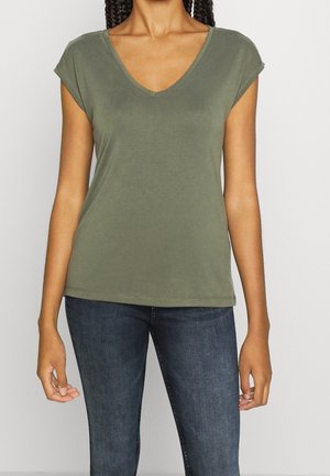 Kvinde iført en olivengrøn V-hals kortærmet skjorte og mørkeblå jeans, stående mod en ensfarvet lys baggrund.