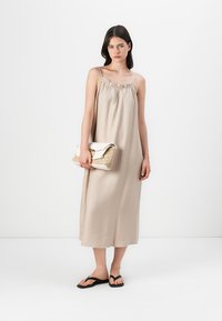 Svijetložuta maksi haljina s podesivim trakama, naboranim izrezom i lepršavom tkaninom; model drži torbu teksture u bež i bijeloj boji.